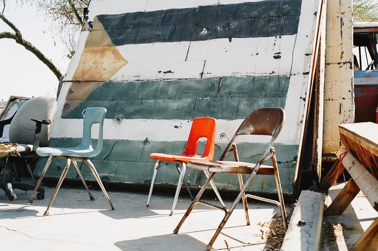 A bright orange folding chair between two muted chairs in a junkyard